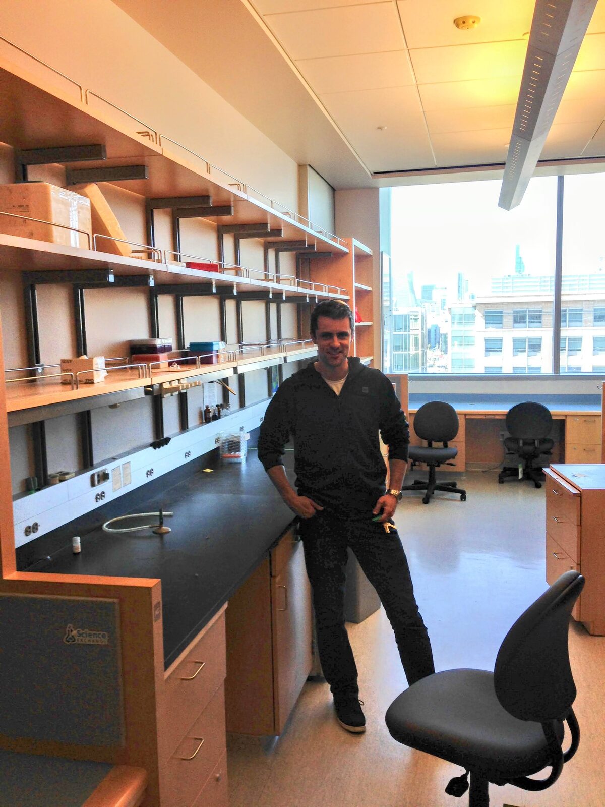 Joe Bondy-Denomy standing in his newly built, still-empty lab at UCSF Mission Bay, beside a long bench with bare shelving and a window looking out over the city